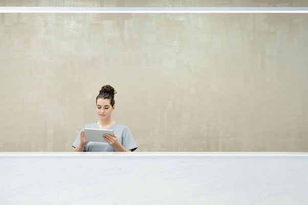 Receptionist in grey scrubs reading a tablet behind a white front desk