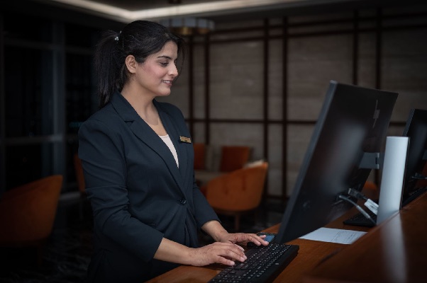 Smiling receptionist in a dark blazer typing at a computer behind a hotel front desk
