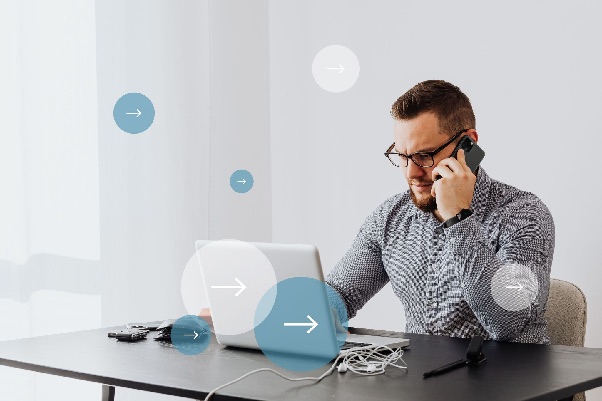 Bearded man in glasses talking on a mobile phone while working on a laptop at a desk, with arrow icons overlaid
