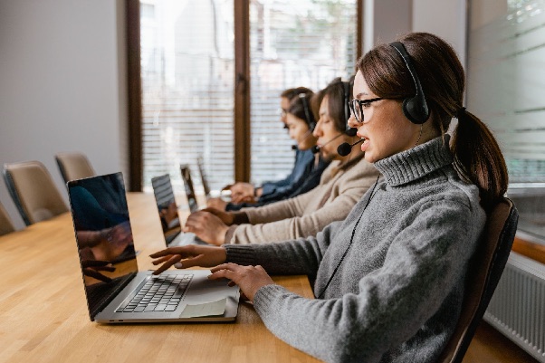 Row of call agents in headsets typing on laptops at a long wooden table by a window