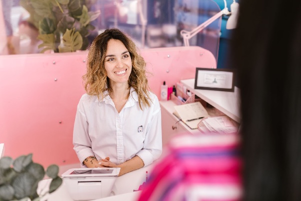 Smiling receptionist in a white shirt greets a visitor from behind a front desk with a pink partition