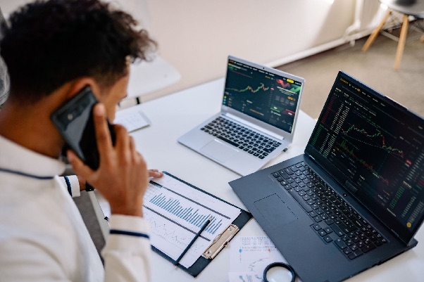 Man on a mobile phone at a desk with two laptops showing market charts and a printed bar chart on a clipboard
